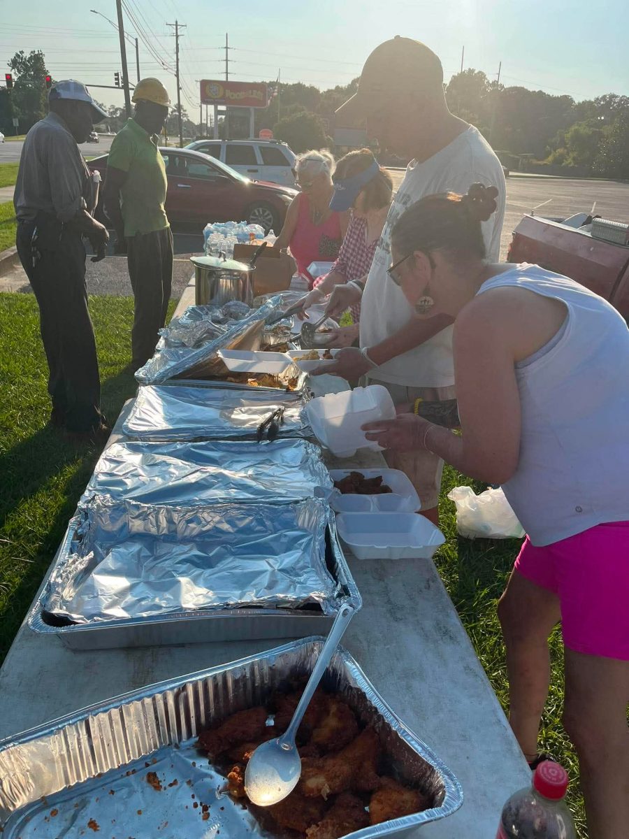 A serving line of foil trays at a Street Feed with volunteers plating fried chicken and sides